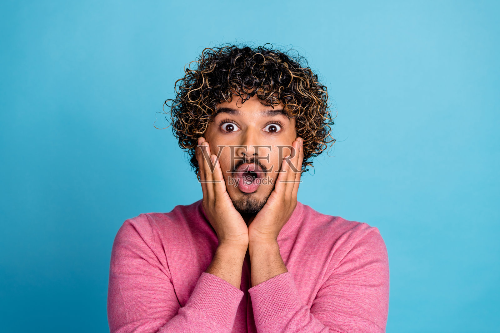 年轻时尚的男士穿着粉色毛衣，卷发，惊讶地双手托腮，背景为对比鲜明的蓝色。照片摄影图片