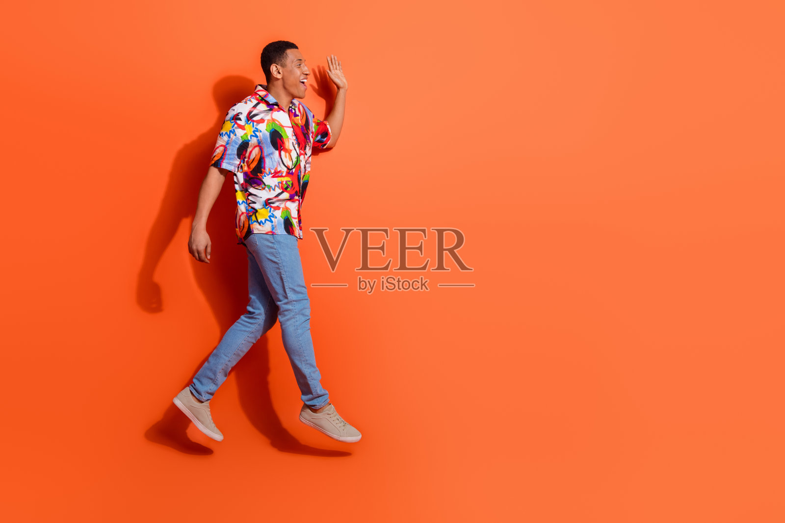 穿着彩色印花衬衫的年轻非裔美国男子在鲜艳的橙色背景下快乐地行走照片摄影图片