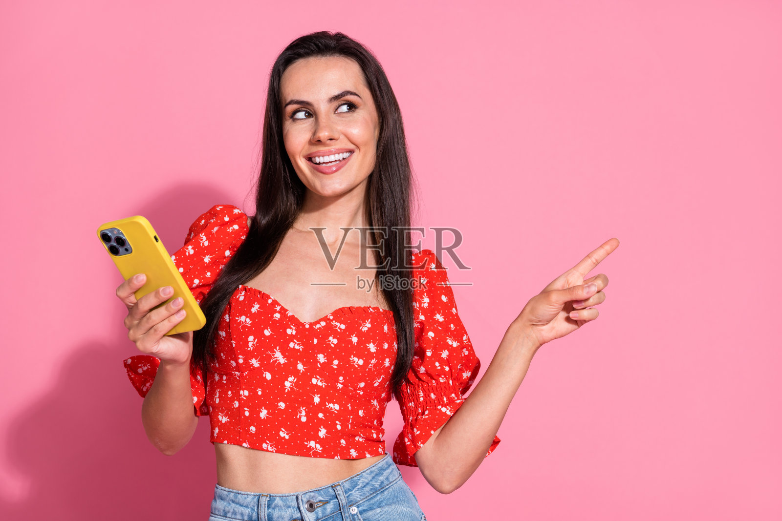 年轻微笑女士穿着明亮红色时尚上衣，展示网站智能手机电子购物功能，背景为粉色。照片摄影图片