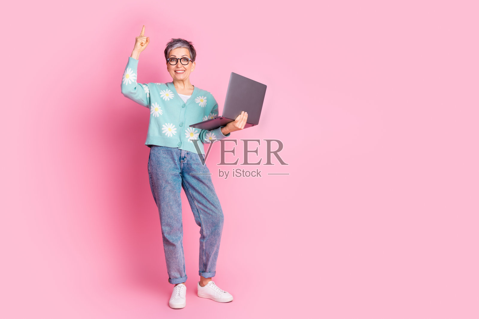 一位老年女性手持笔记本电脑，指向上方，站在粉色背景前。照片摄影图片