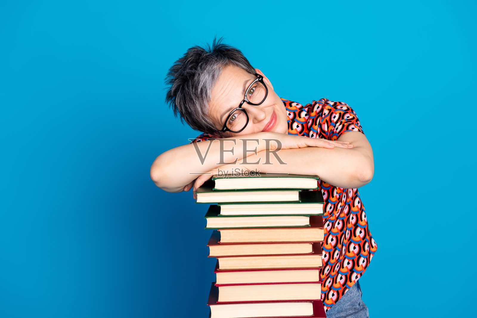 迷人的戴眼镜女性倚靠在一堆书籍上，背景为明亮的蓝色，展现出休闲和生活方式。照片摄影图片