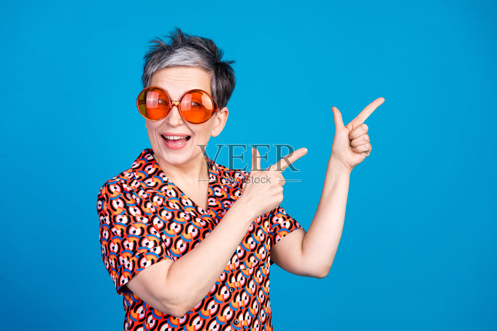 快乐的老年女性戴着现代眼镜，指向蓝色背景的一侧照片摄影图片