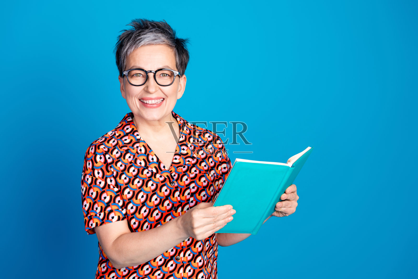微笑的成熟的女性戴着眼镜，手持一本书，背景为时尚的蓝色，穿着图案衬衫。照片摄影图片
