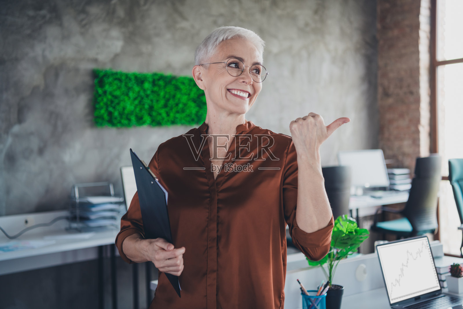 专业成熟的女性企业领导者的照片，指向空白空间，穿着棕色衬衫，在明亮舒适的办公室内共事。照片摄影图片