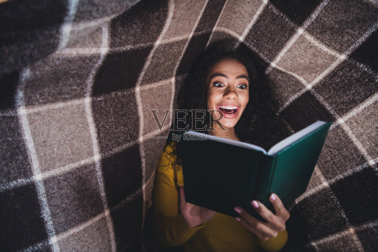 年轻女性在室内享受一本引人入胜的书，展现出愉悦和惊讶的表情，坐在温馨的环境中，裹着毯子。照片摄影图片