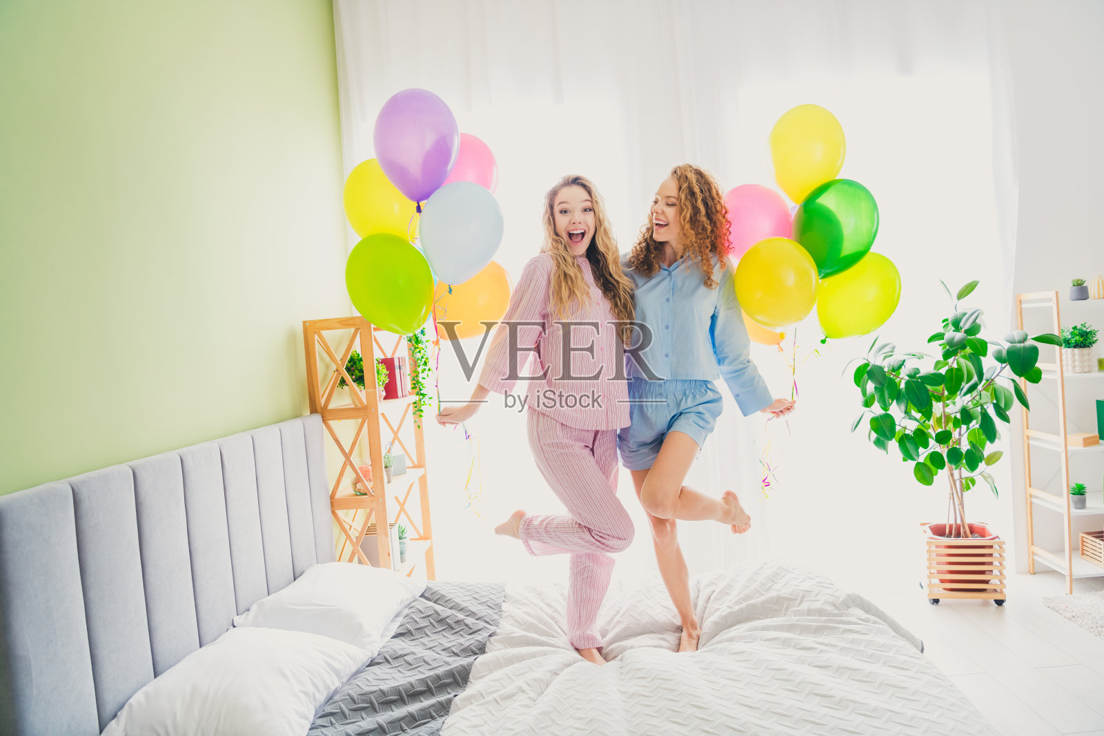 两位可爱年轻女孩在现代舒适明亮的卧室里穿着睡衣庆祝生日，享受起床的乐趣。照片摄影图片