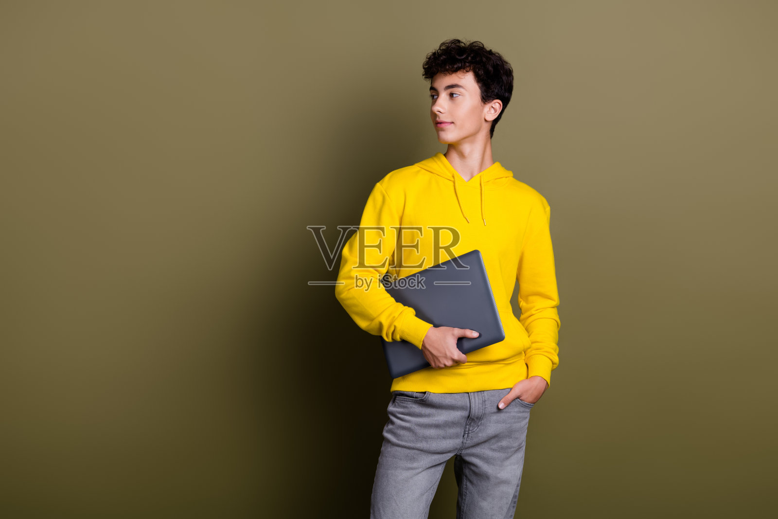 年轻时尚的男子穿着黄色连帽衫，手持笔记本电脑，背景为卡其色，外观休闲。照片摄影图片