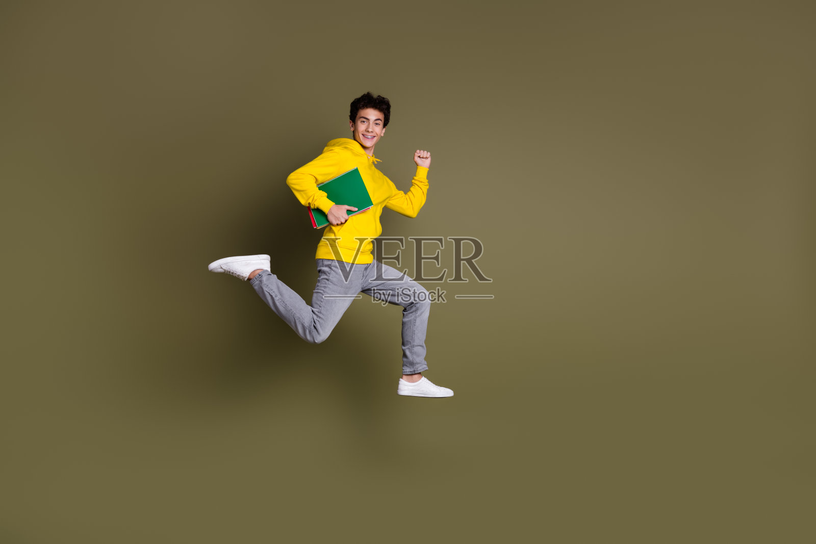 穿着休闲的年轻学生在卡其色背景上欢快地跳跃，手中拿着书籍。照片摄影图片