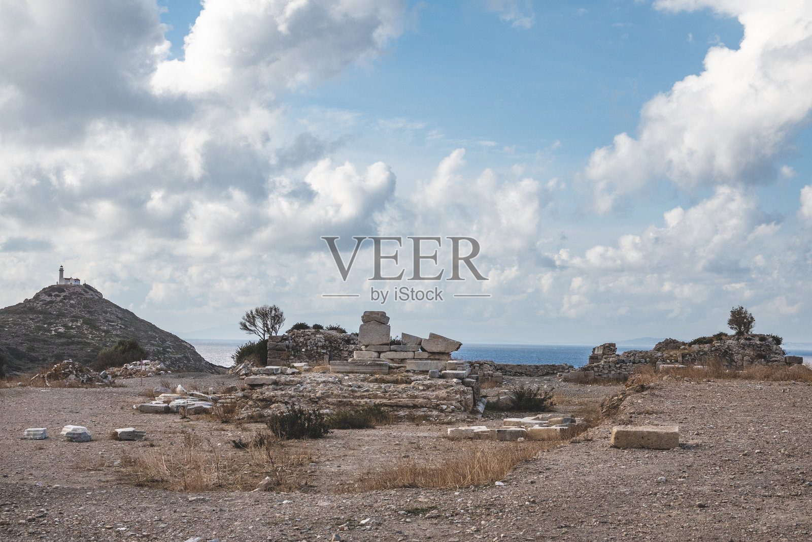 一个古老的考古遗址，靠近海岸，散落着废墟，背景中有宁静的海洋和山丘上的灯塔，天空部分多云。照片摄影图片