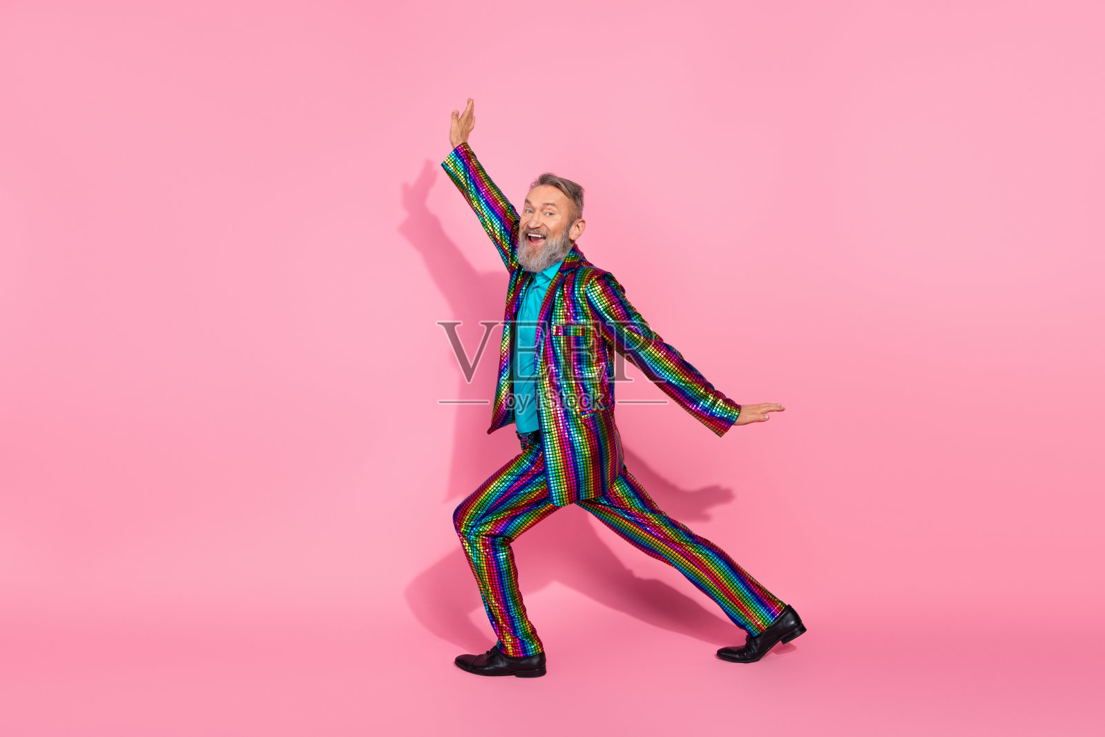 充满魅力的老年男子穿着鲜艳的彩虹西装，在粉色背景前快乐地跳舞照片摄影图片