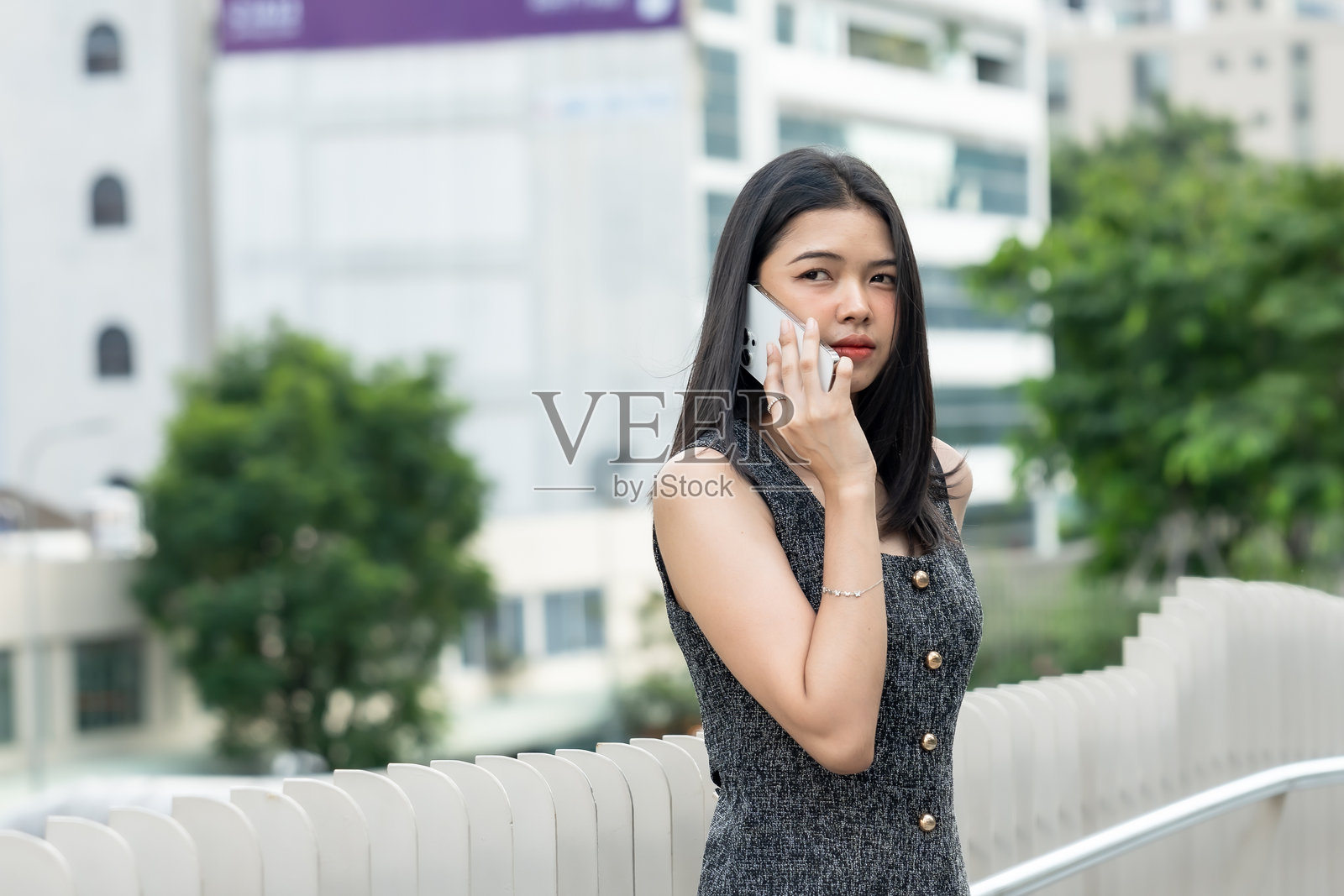 美丽的亚洲办公室工作人员在商业区走动。年轻女性站在城市护栏旁，专注地在智能手机上通话，周围是现代建筑的城市环境。照片摄影图片