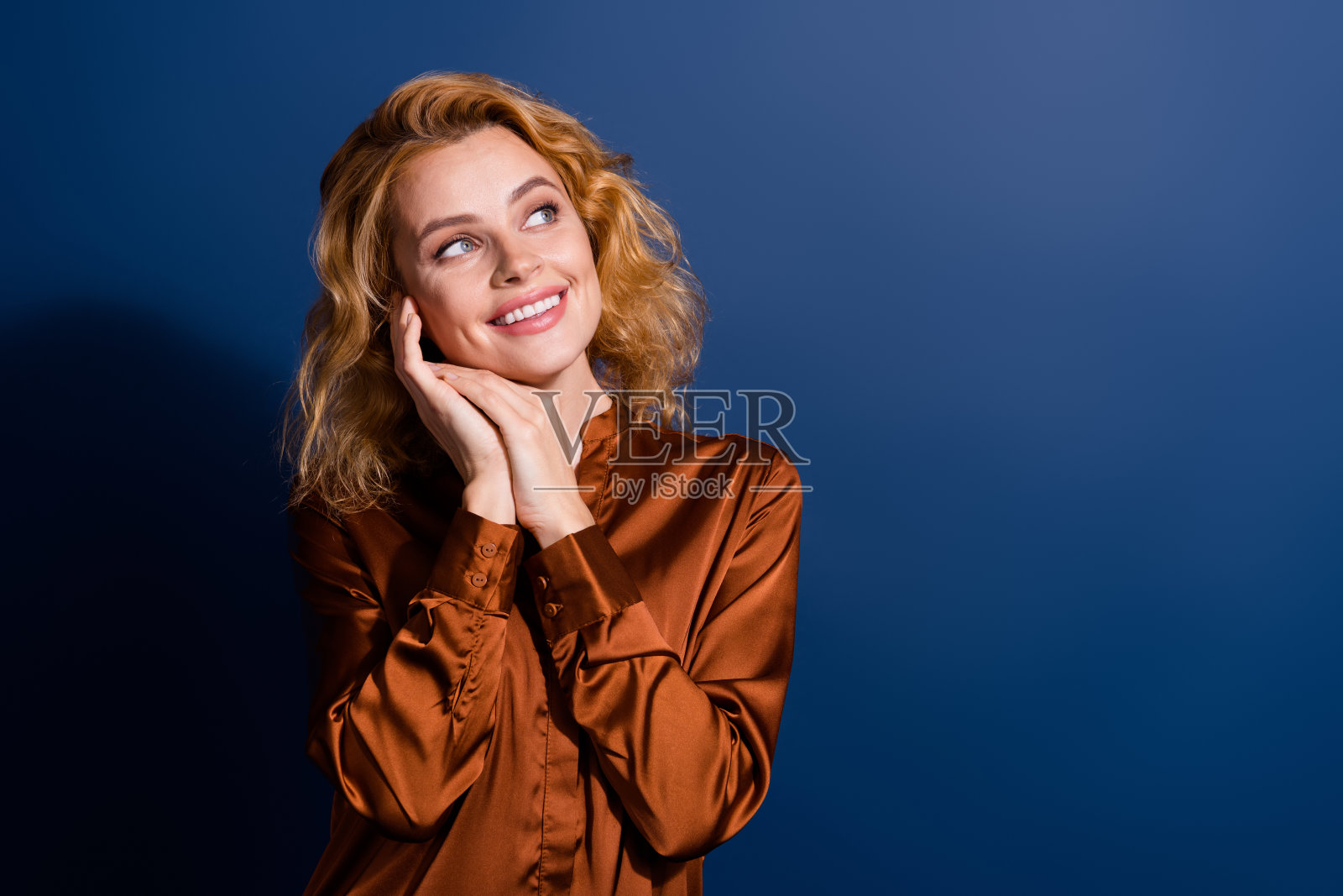 心情愉悦的梦幻女性，波浪卷发，穿着丝质衬衫，双臂托腮，凝视空旷的空间，背景为深蓝色。照片摄影图片