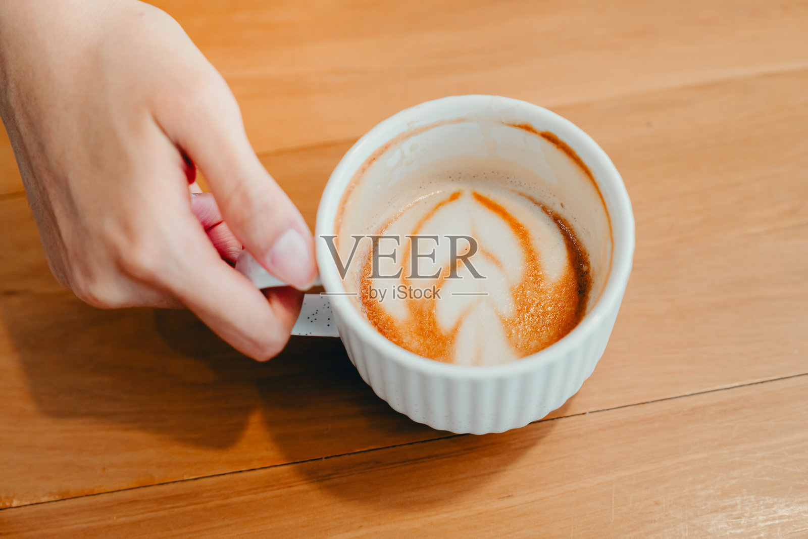 手握白色陶瓷杯，杯中有拉花，放在木质桌面上照片摄影图片