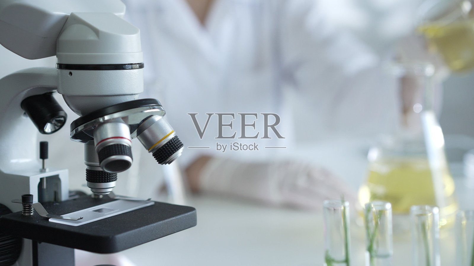 一位科学家穿着实验室外套和白色防护手套，正在实验室中将黄色油状液体从一个烧杯倒入另一个烧杯，靠近显微镜，特写镜头。医学与科学概念。照片摄影图片