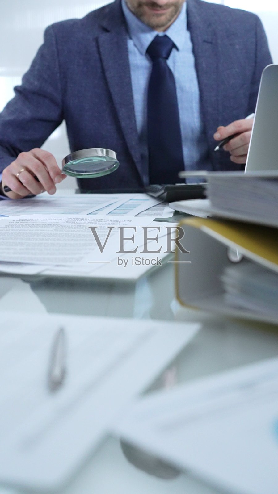 身穿蓝色西装的商人在专业办公室环境中使用放大镜分析财务报告，并使用计算器。低光照，特写竖视图。商业人士，审计。照片摄影图片