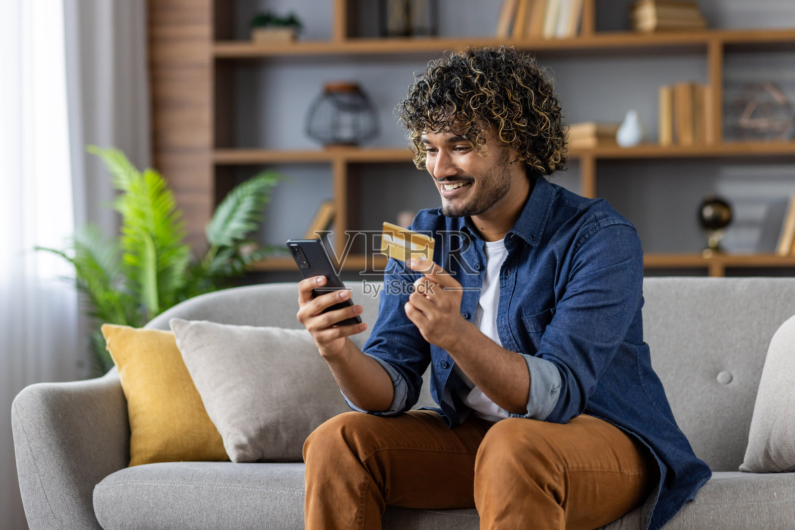 微笑的男人坐在沙发上用智能手机和信用卡进行在线购物照片摄影图片