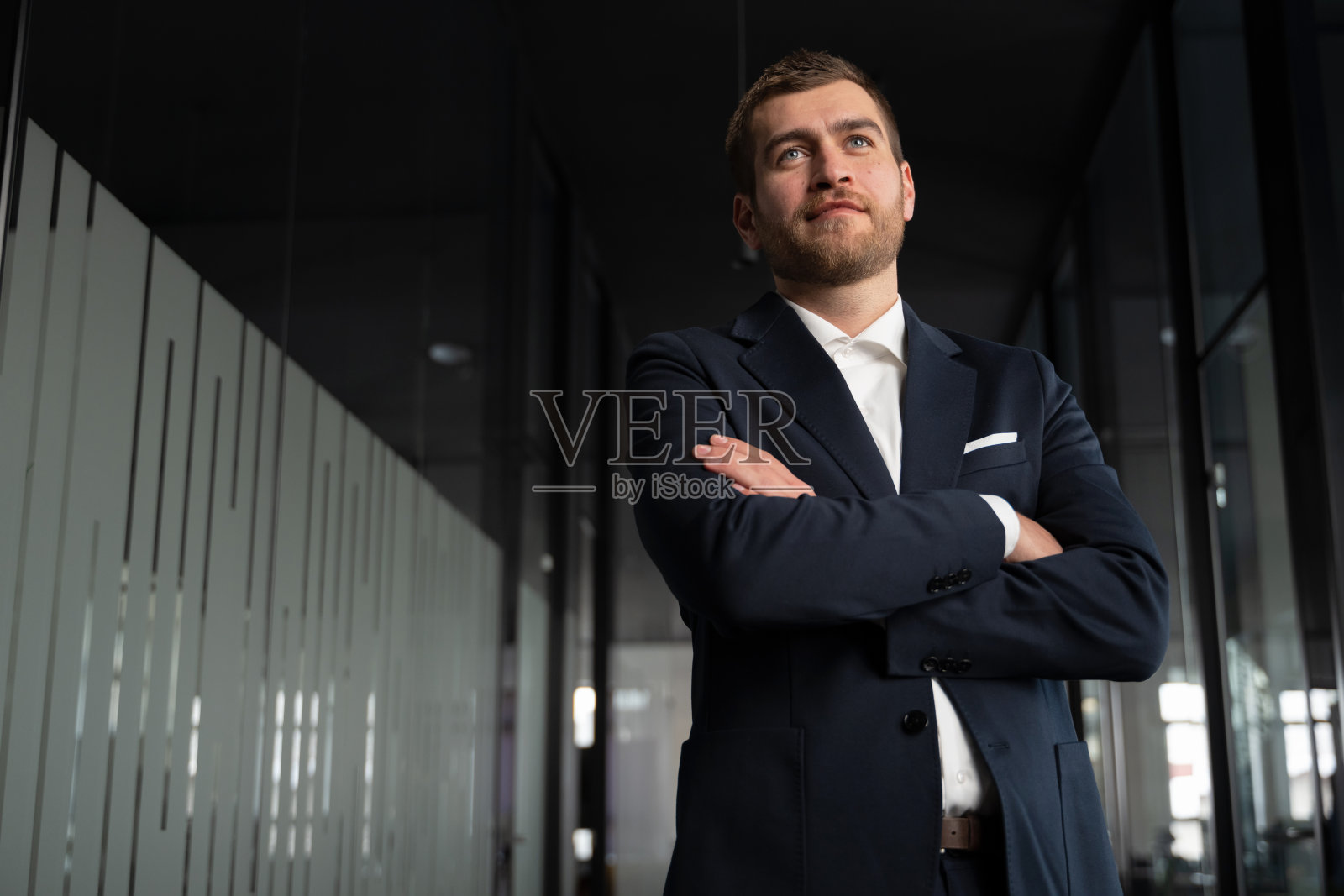 自信的商人、执行官或企业家穿着海军蓝西装，双臂交叉站在现代办公室走廊中。领导力、成功、企业自信和职业抱负。照片摄影图片