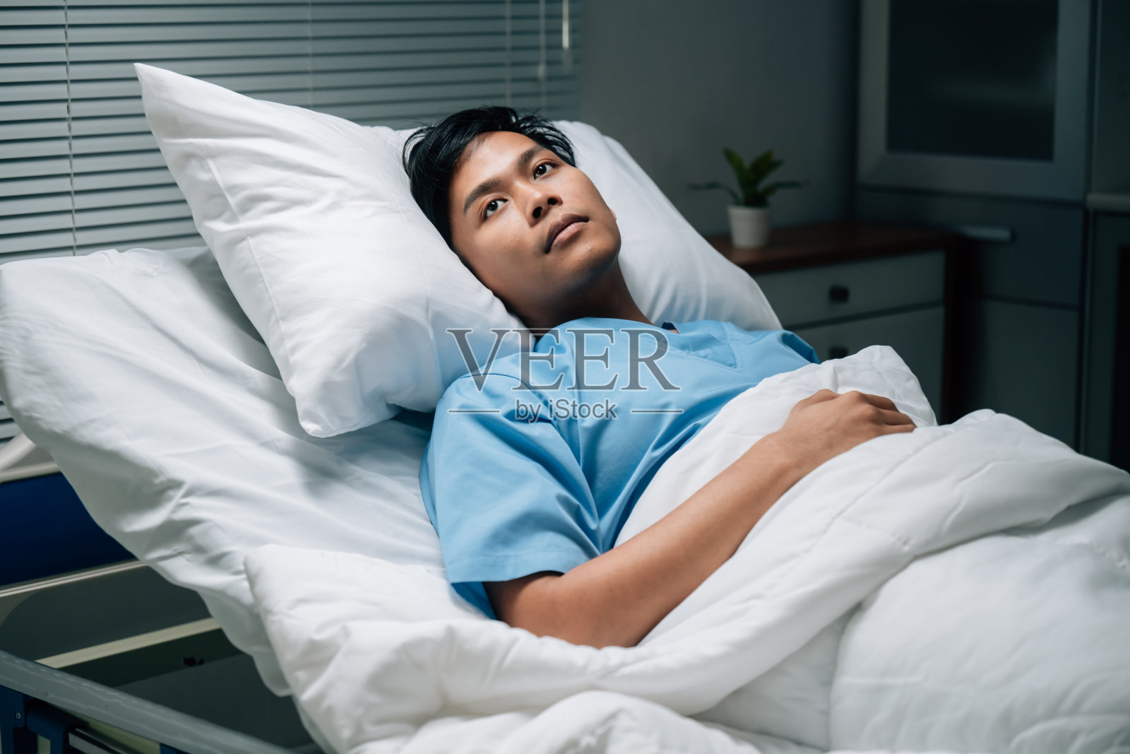 医院病人面露担忧，躺在床上，穿着医疗袍，在昏暗的环境中展现脆弱和焦虑，适合医疗相关的图像。照片摄影图片