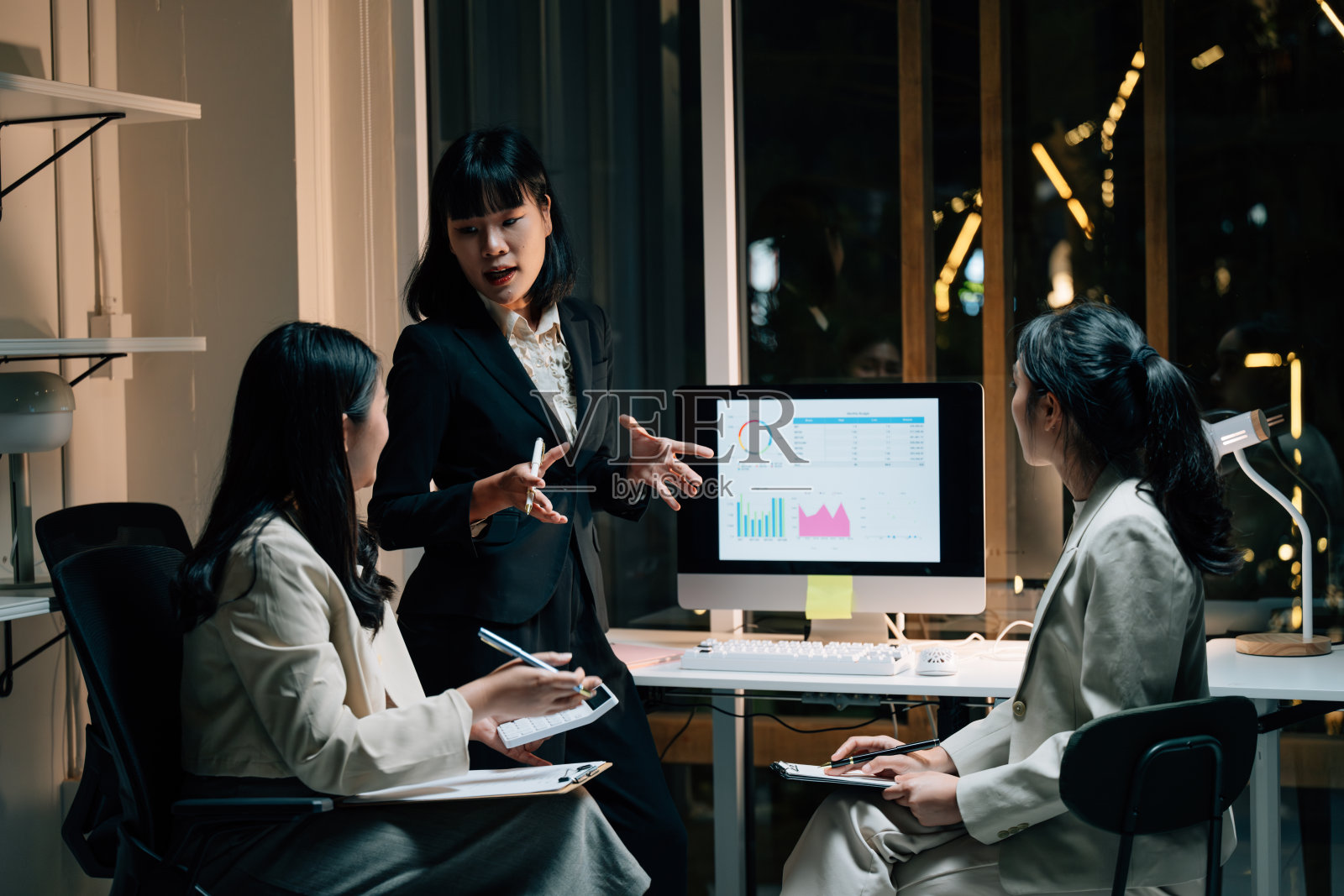 三位年轻的亚洲女商人在现代办公室进行深夜会议，专注地分析电脑屏幕上的图表和数据。照片摄影图片