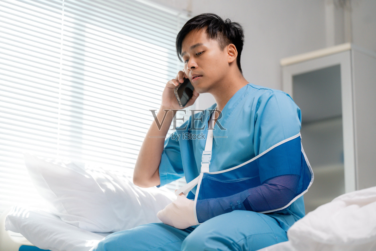 年轻成年患者穿着蓝色手臂吊带，坐在医院病床上，通过手机沟通，正在康复中。照片摄影图片