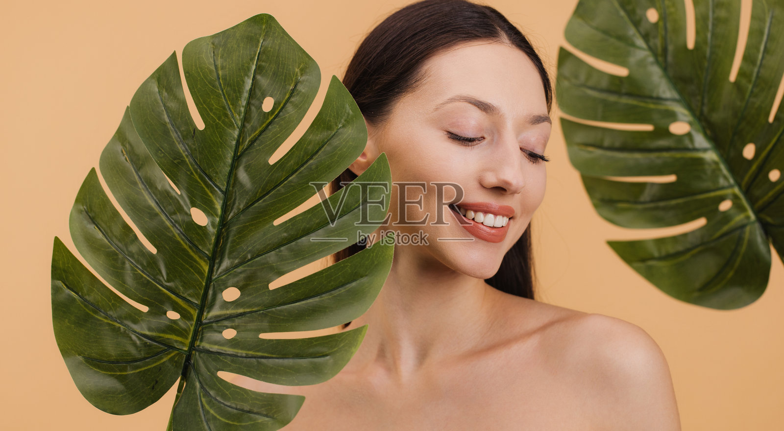 美丽微笑的女性与米色背景上的热带龟背竹叶合影。美容概念。照片摄影图片