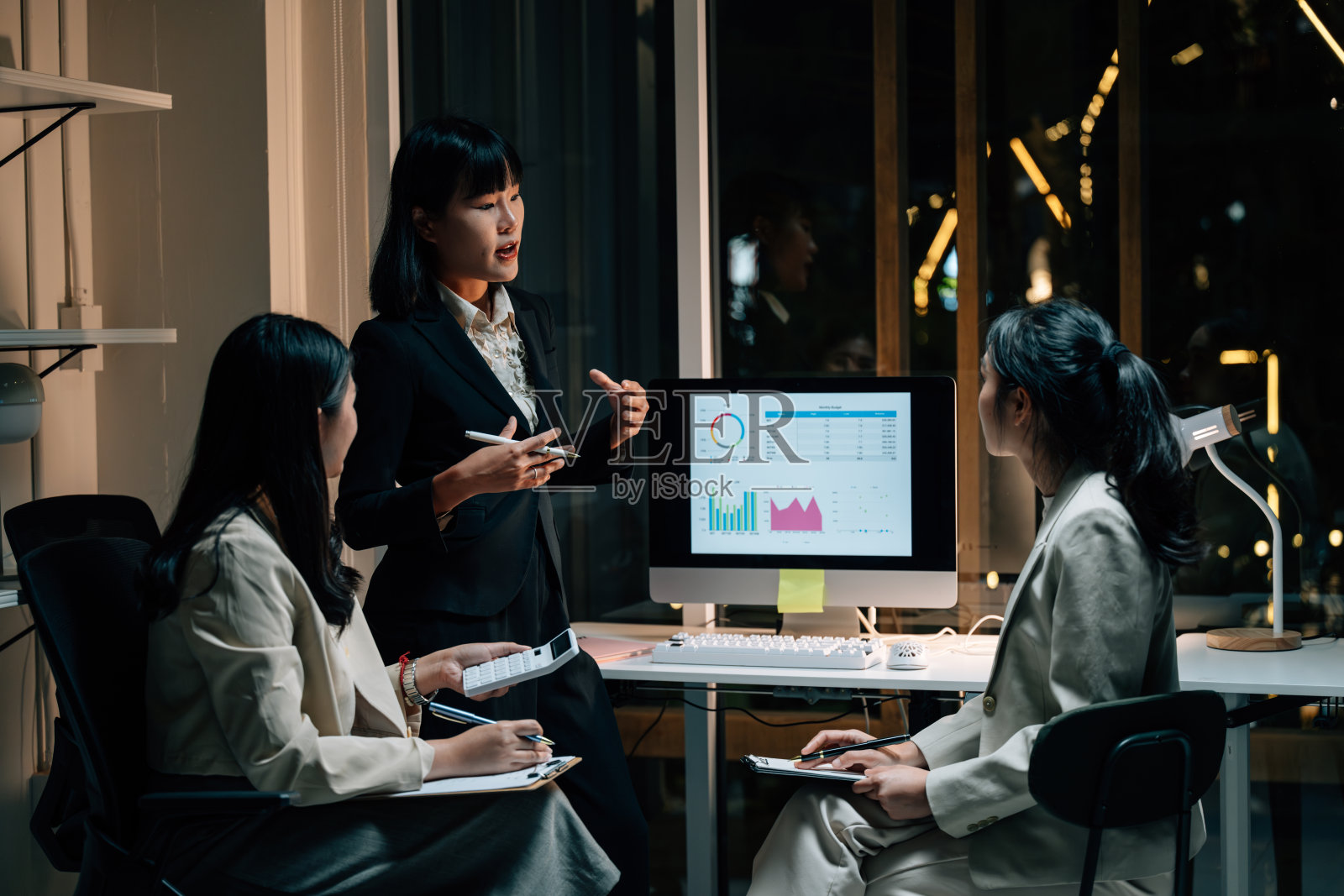 三位年轻的亚洲女商人在办公室熬夜开会，讨论图表并分析电脑屏幕上的财务数据。照片摄影图片