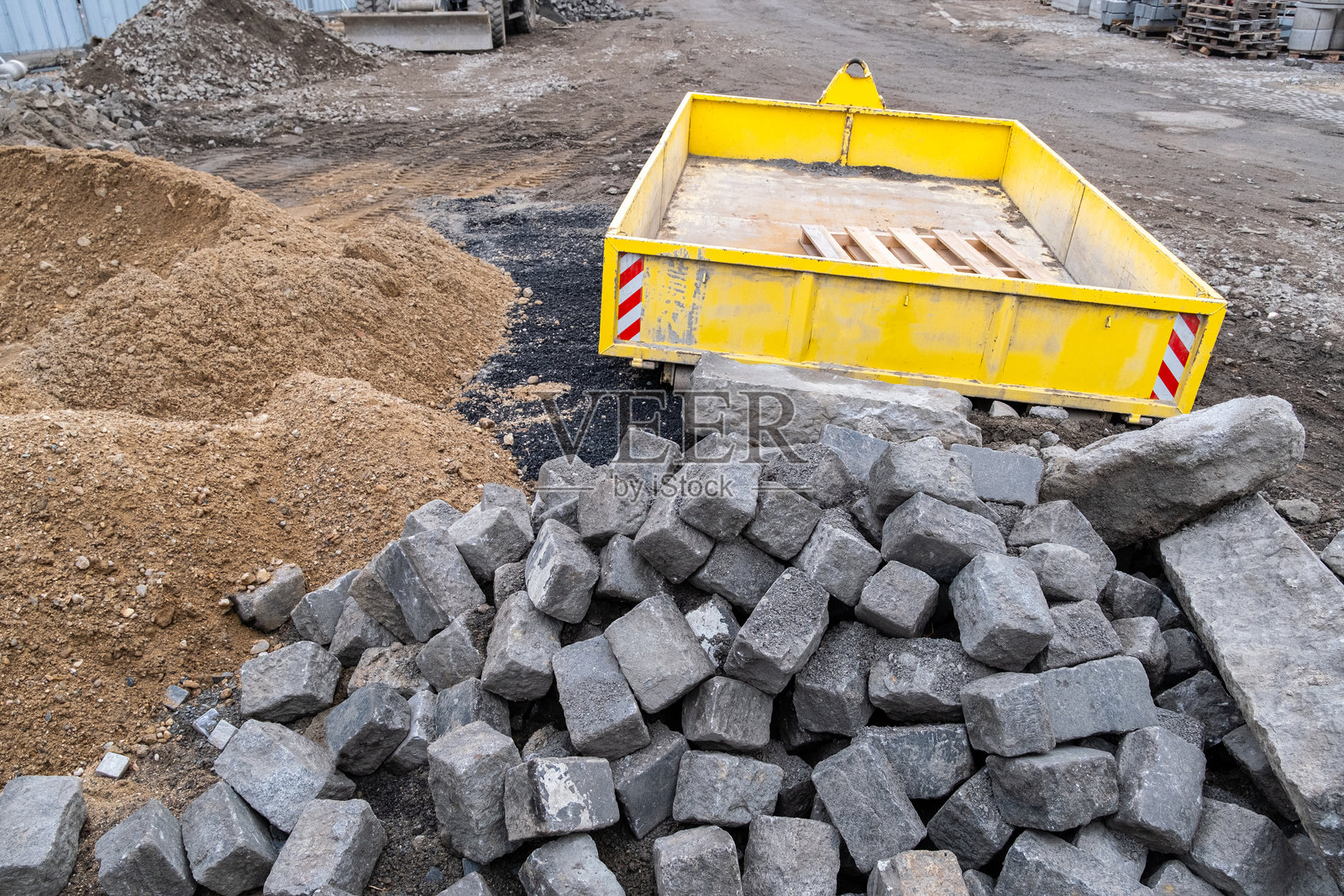 一个建筑工地，附近有堆积的卵石、沙子和碎石，以及一个黄色的废物容器。该区域已为道路施工或基础设施开发做好准备。照片摄影图片