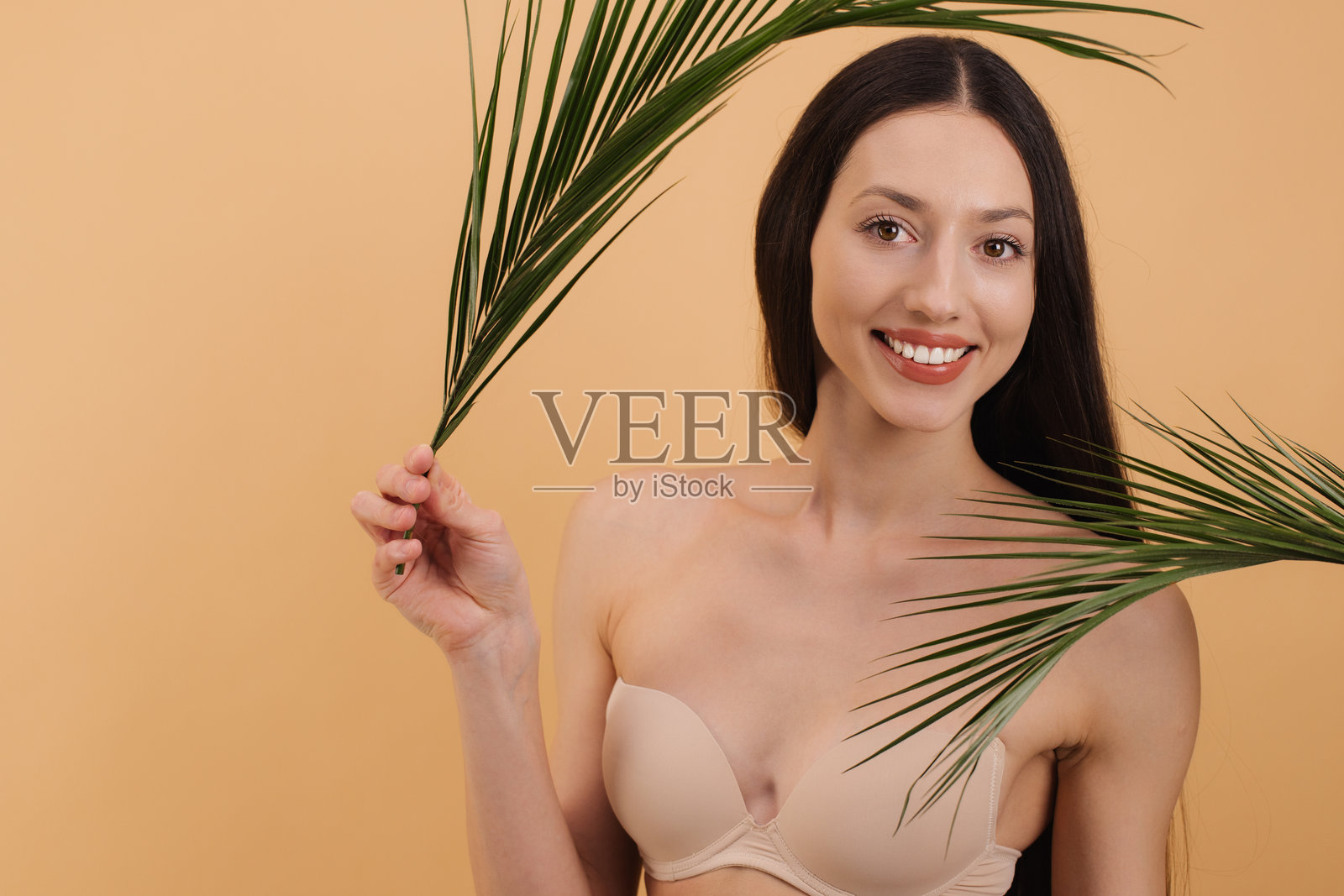 美丽微笑的女性手持热带棕榈叶，摆姿势于米色背景前照片摄影图片