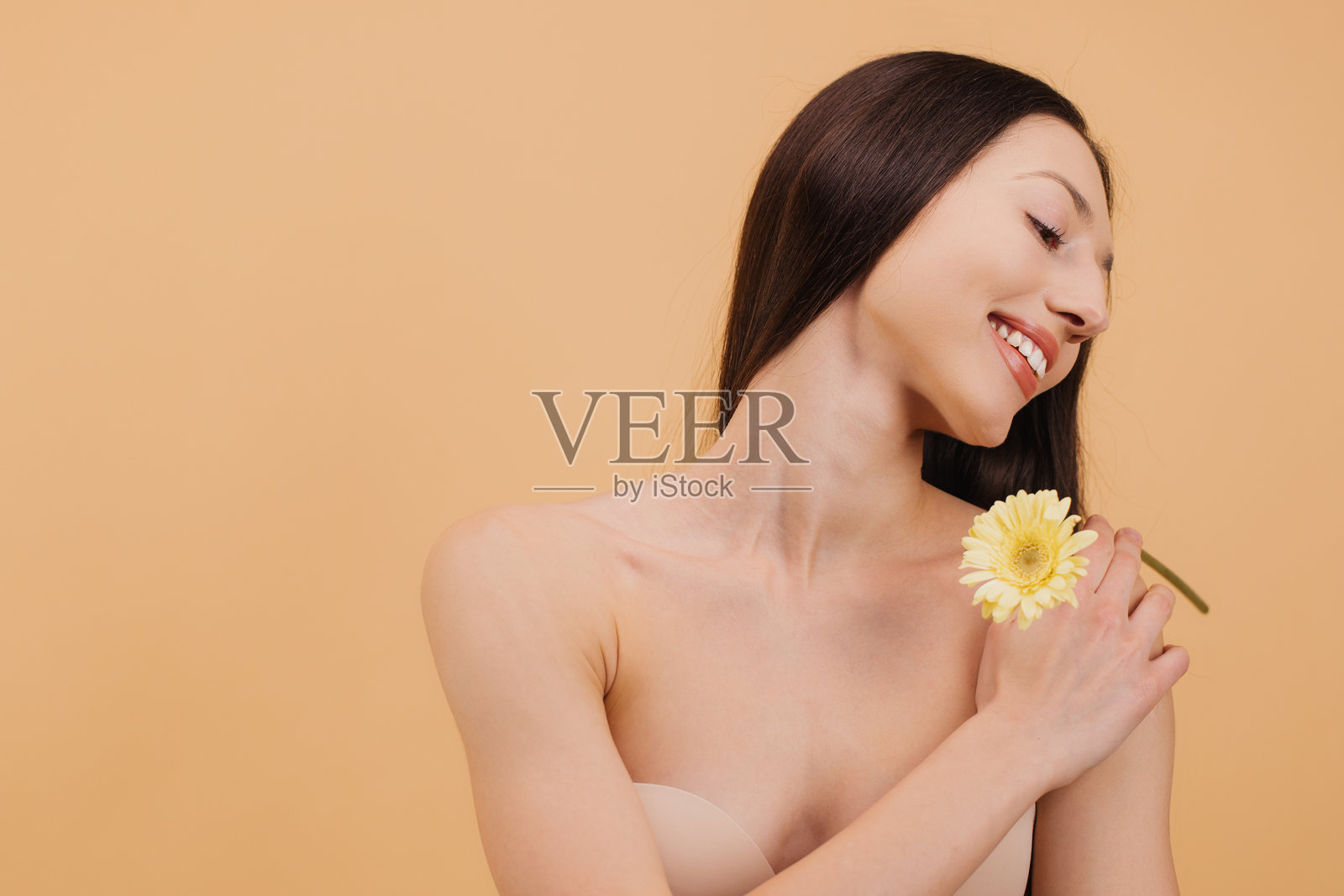 美丽微笑的女性在米色背景下，光裸的肩膀上托着一朵黄色花。照片摄影图片