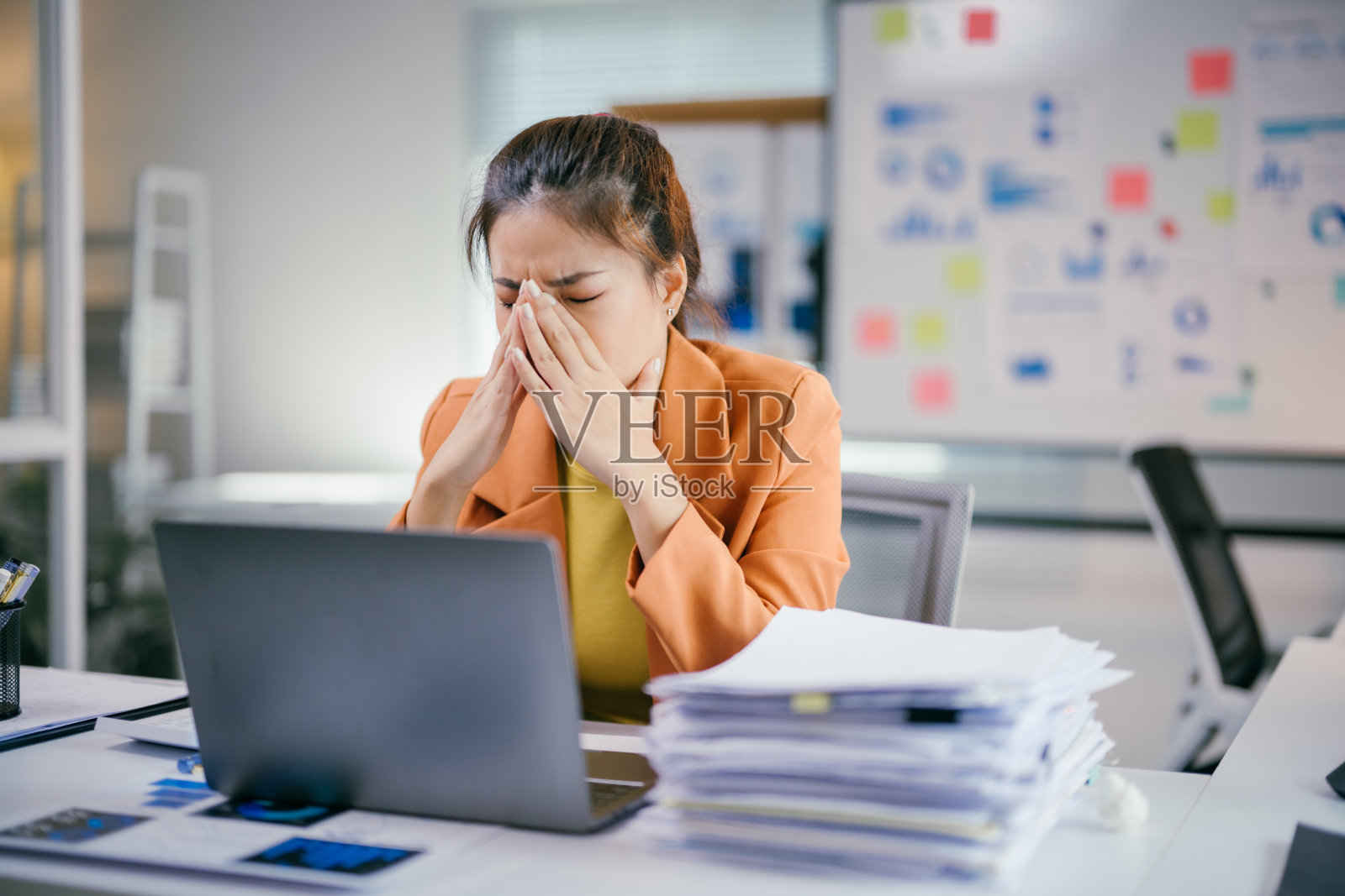 专业的亚洲女商人在拥挤的办公室环境中感受到强烈的压力，双手按压额头，坐在工作空间内。照片摄影图片