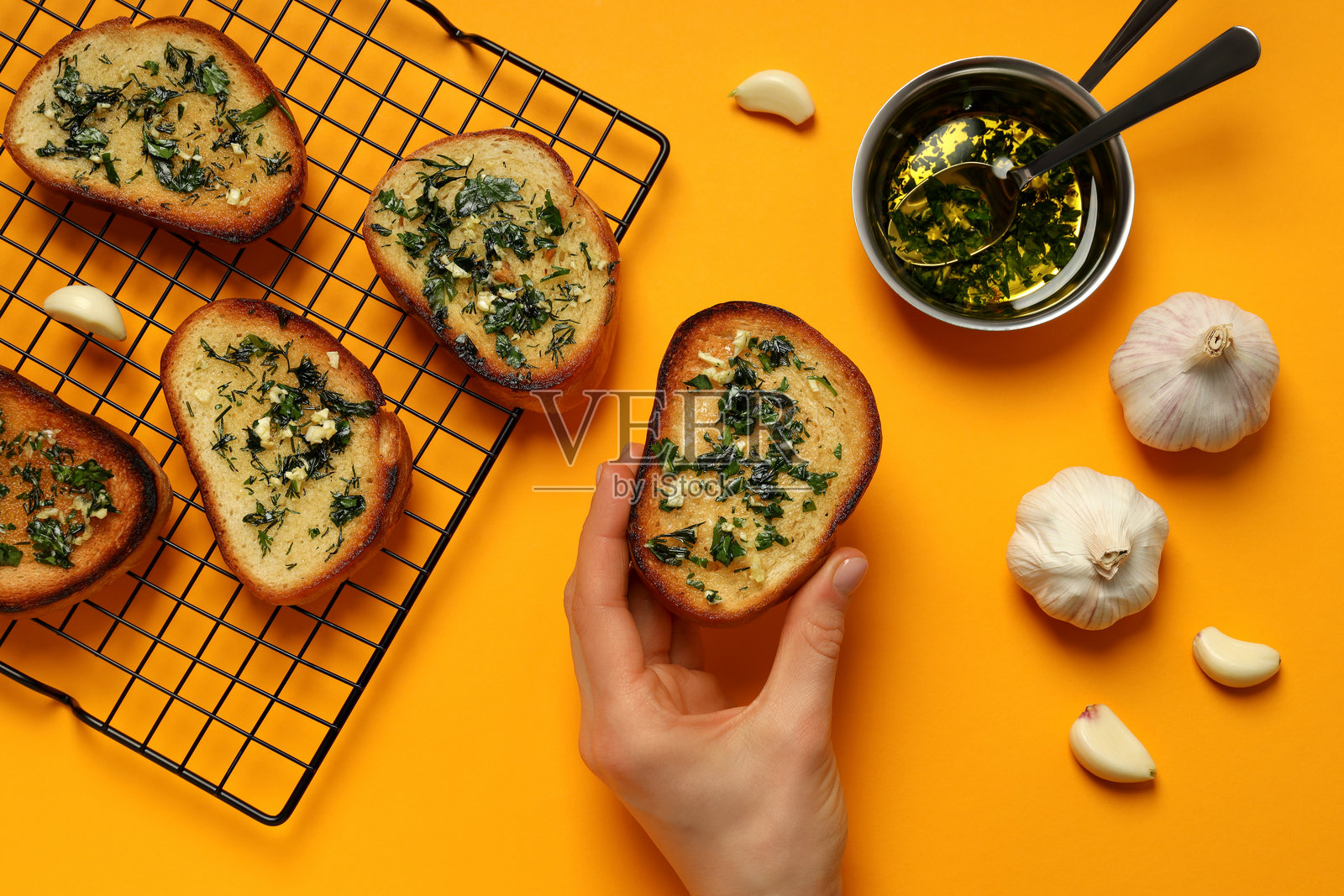 手中握着的蒜蓉面包片与网格上的蒜蓉面包，旁边有大蒜和装有油的碗，黄色背景，俯视图照片摄影图片
