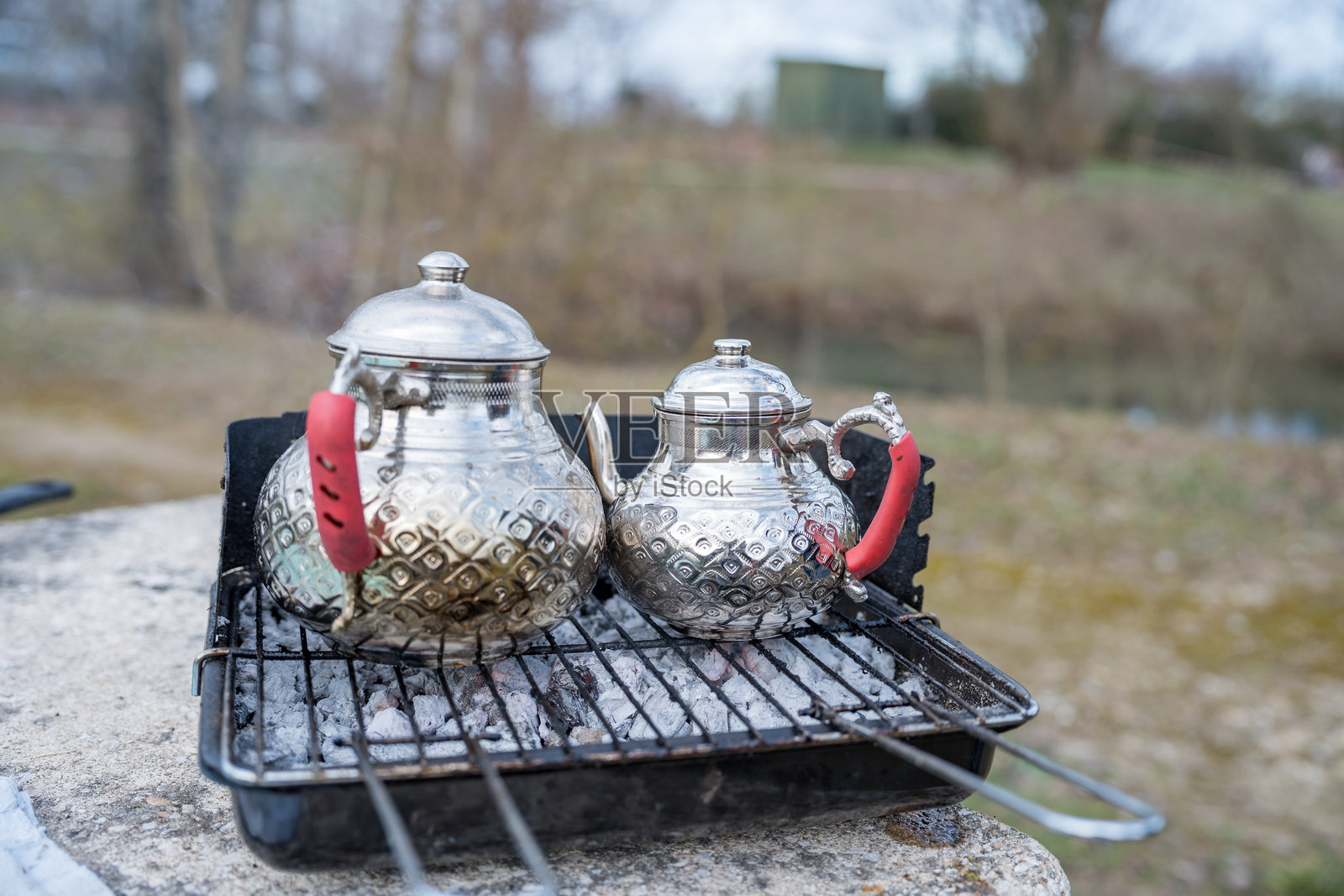 银色茶壶在河边的烤架上轻轻冒着蒸汽，朋友们在宁静的下午聚会。照片摄影图片