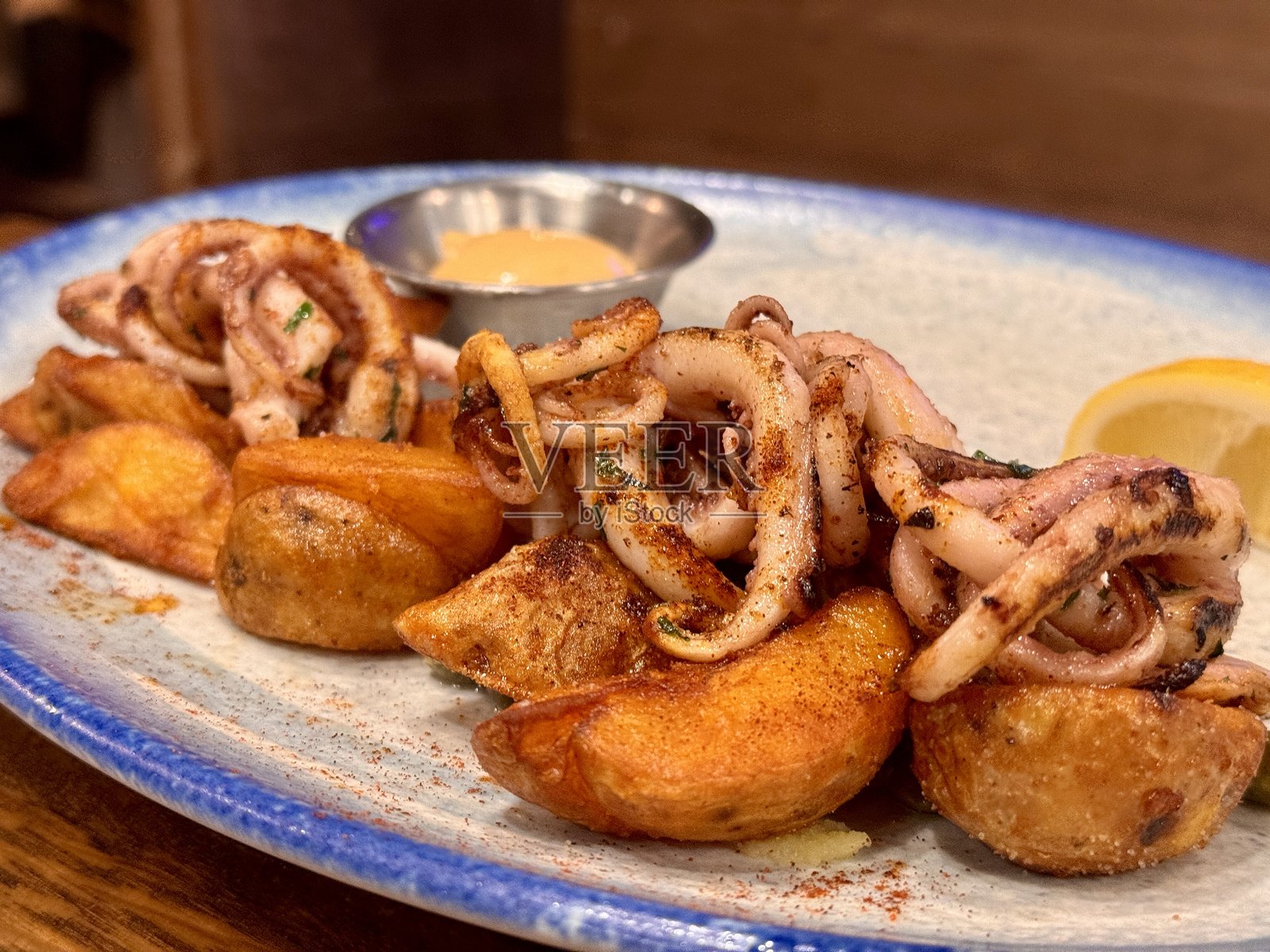 白色盘子里盛着油炸章鱼、土豆和酱汁，放在海鲜餐厅的木桌上。特写。照片摄影图片