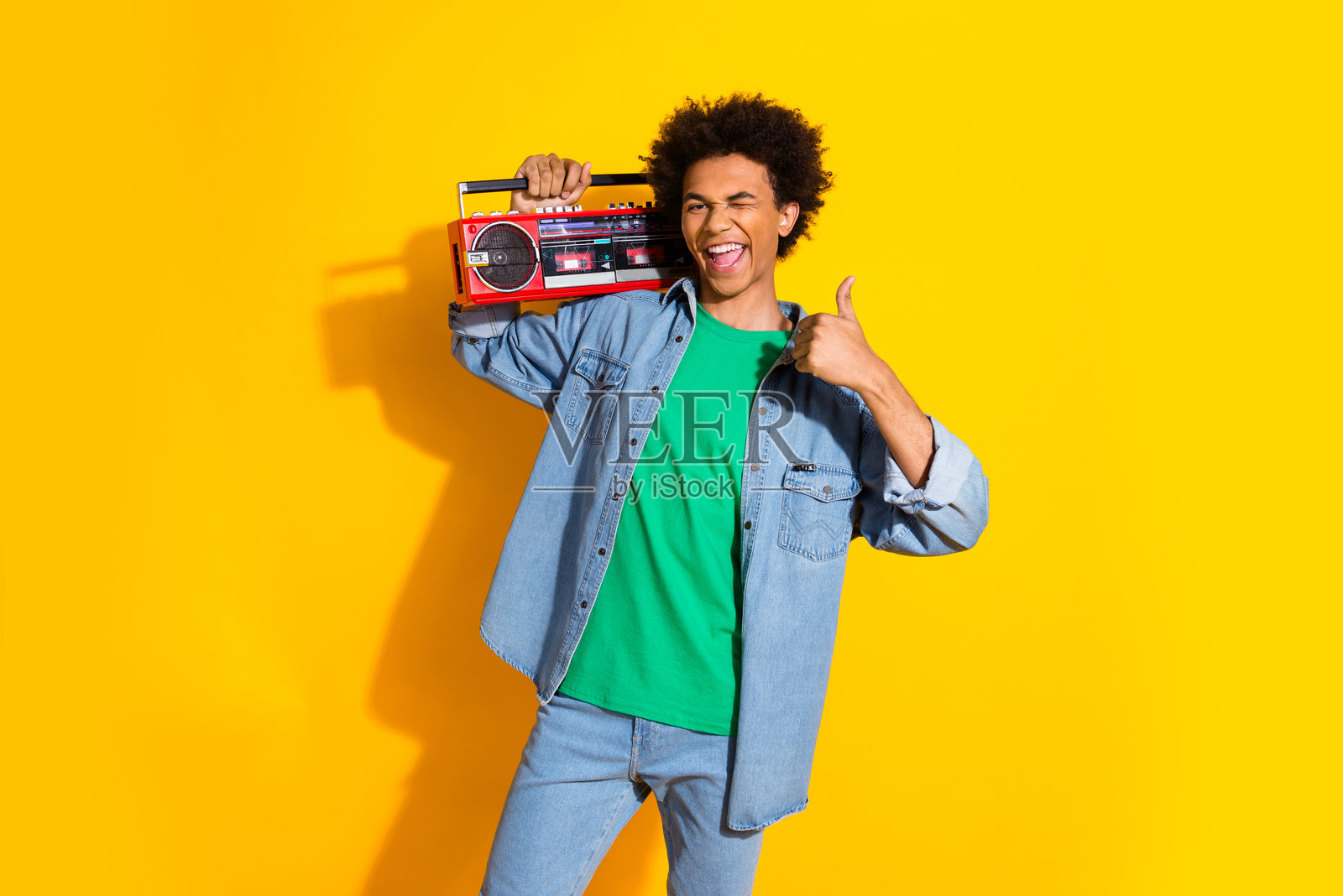 年轻帅气的小伙子眨眼竖起大拇指，手持音响，穿着牛仔衬衫，背景为黄色。照片摄影图片