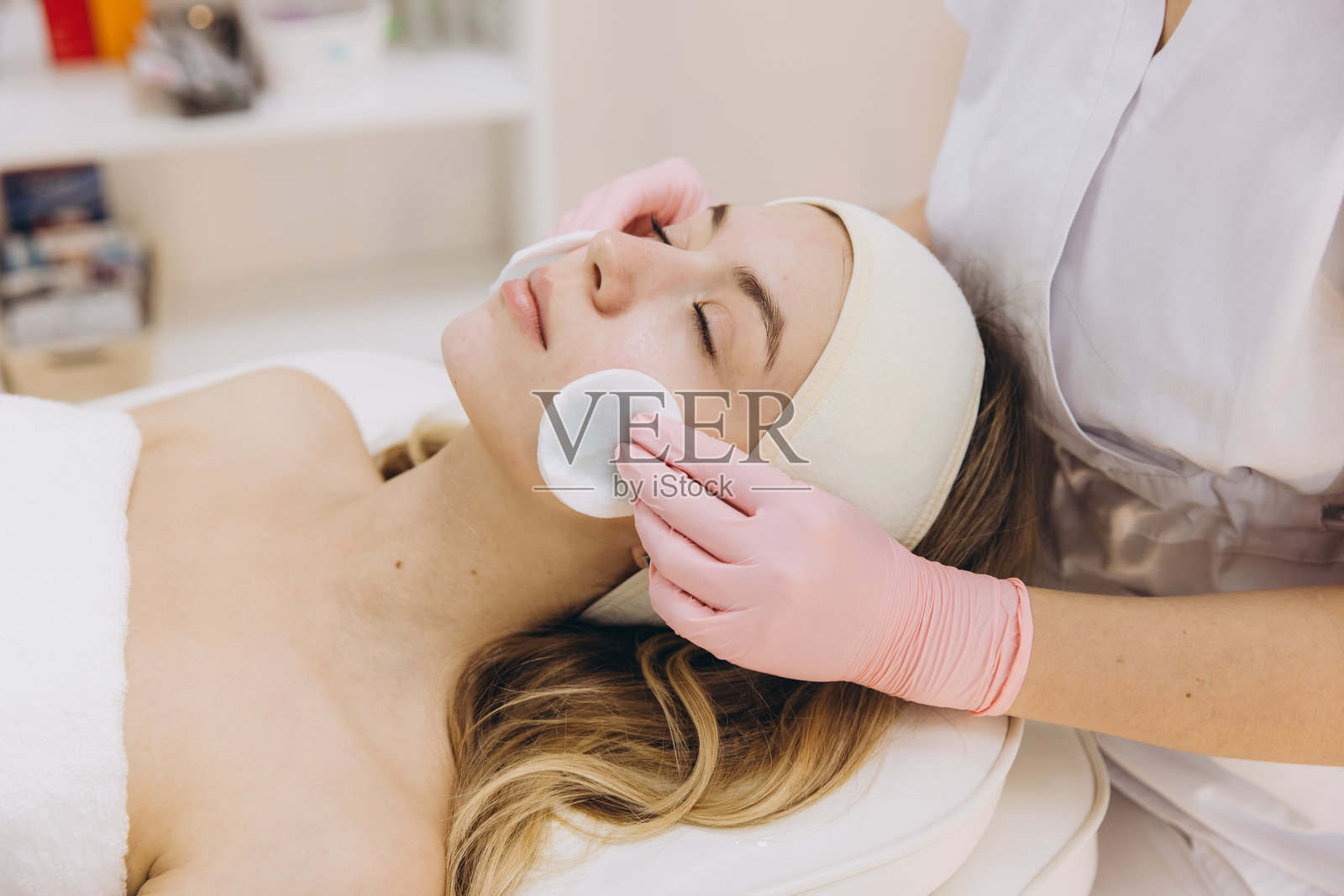 专业美容师戴着粉色手套，正在水疗沙龙为金发客户温柔地用棉垫清洁面部。照片摄影图片