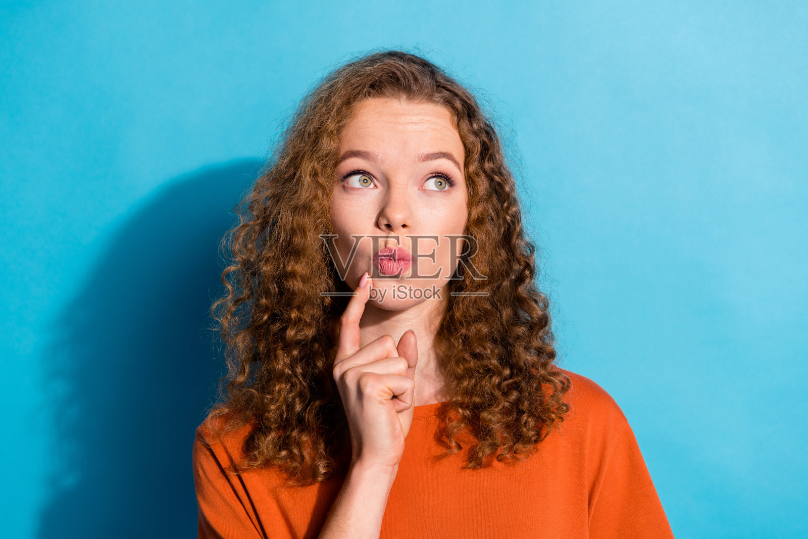 青春美丽的有趣女孩，手托下巴，神情恍惚，波浪卷发，穿着橙色T恤，背景为蓝色。照片摄影图片