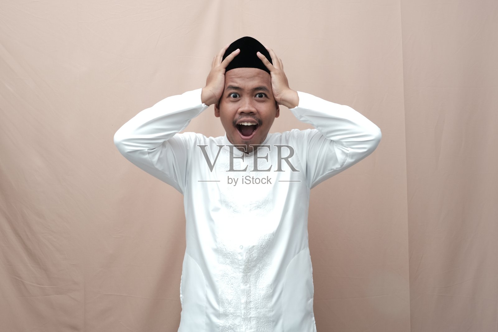 震惊的亚洲穆斯林男子，穿着伊斯兰服装，嘴巴张开，双手放在头上照片摄影图片