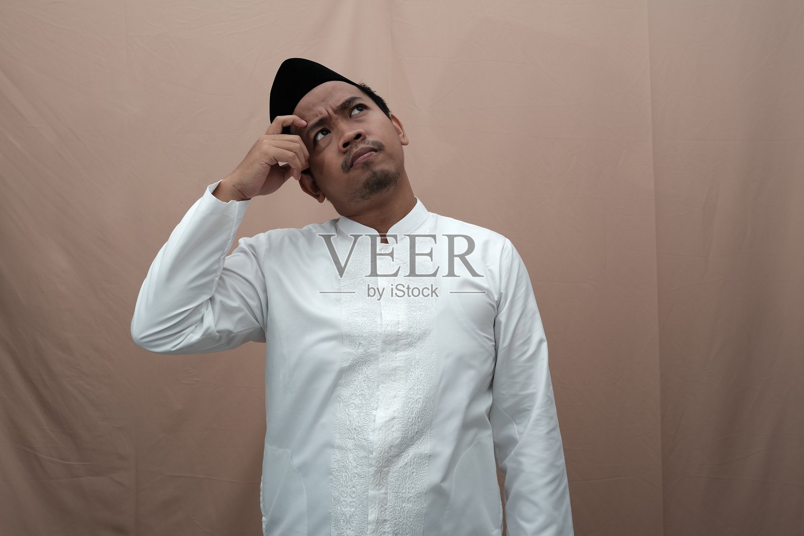 深思的亚洲穆斯林男子，身穿传统白色服装，手托下巴，仰望着天空。照片摄影图片