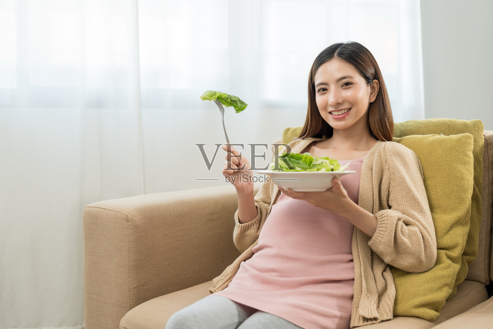 快乐的孕妇吃着蔬菜沙拉，微笑着看向镜头，体现了健康母亲关注自身健康、滋养胎儿并期待分娩的概念。照片摄影图片