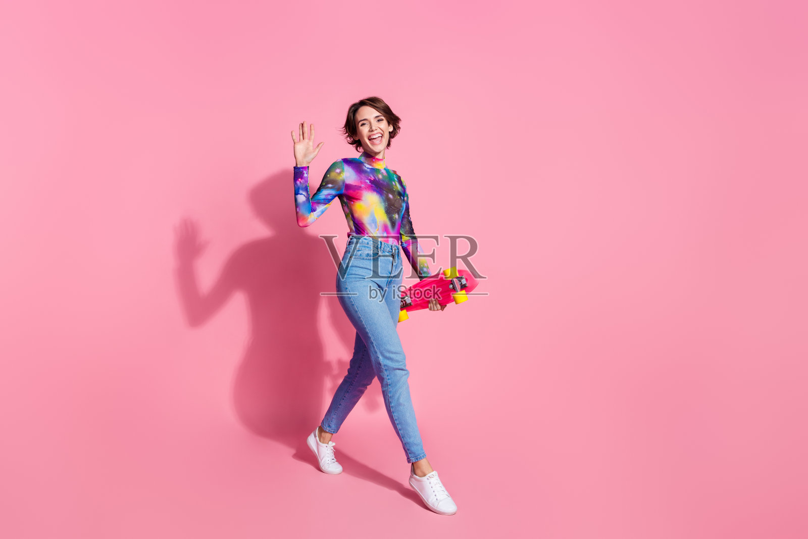 全身肖像的漂亮女孩，手持滑板，挥手打招呼，穿着套头衫，背景为粉色。照片摄影图片