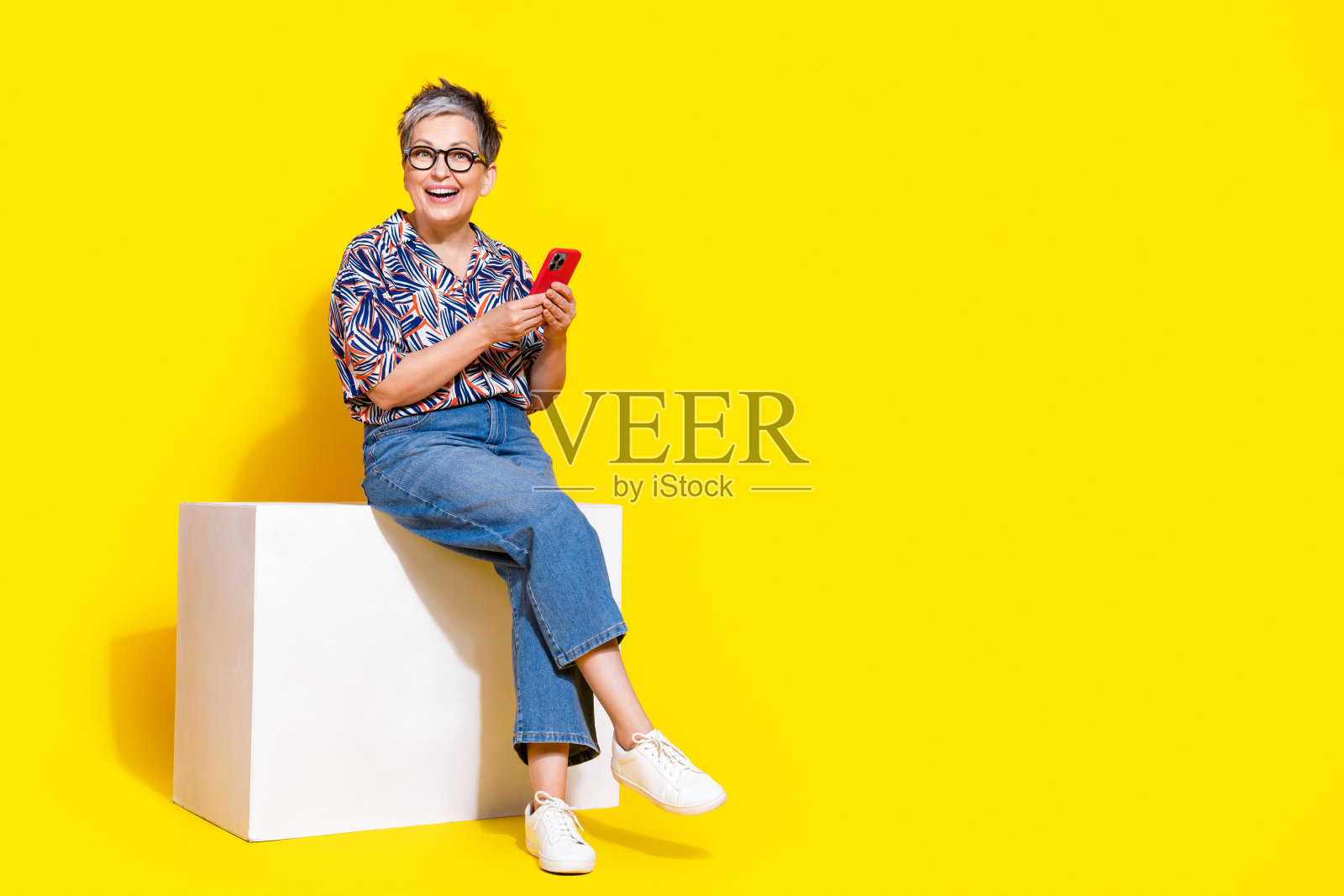 全身照，迷人的老年女士坐在立方体上，手持设备，穿着时尚的彩色服装，背景为黄色。照片摄影图片