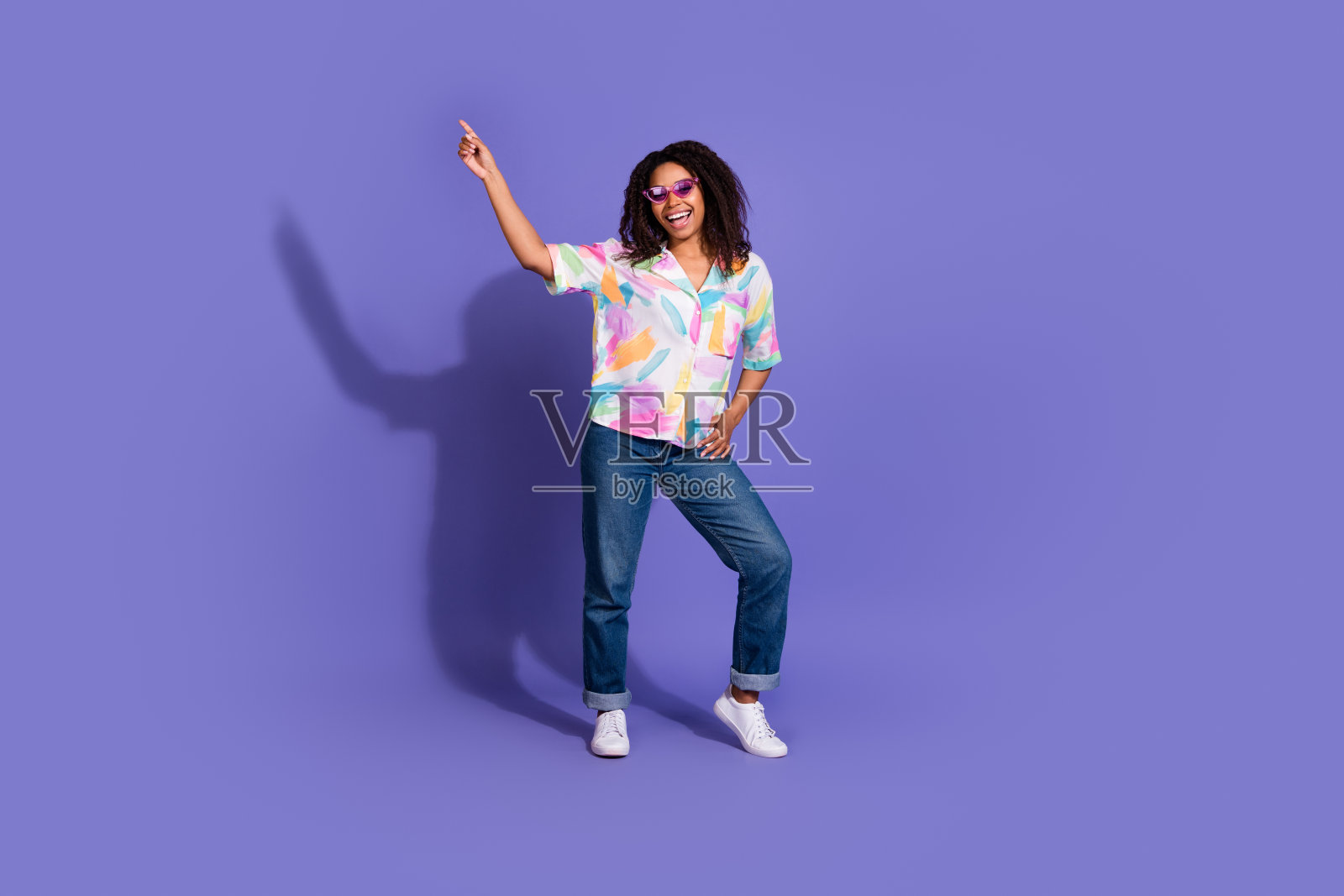 年轻女性穿着鲜艳的衬衫，在紫色背景下快乐地跳舞，戴着太阳镜和牛仔裤，展现出休闲风格和魅力。照片摄影图片