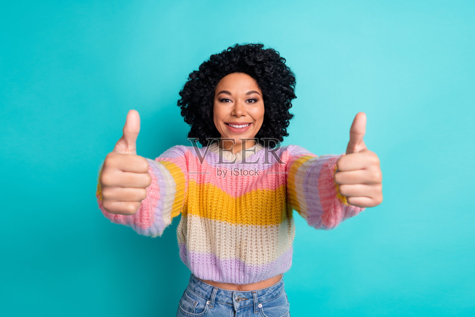 穿着鲜艳多彩毛衣的快乐年轻女性在青色背景前竖起大拇指。照片摄影图片