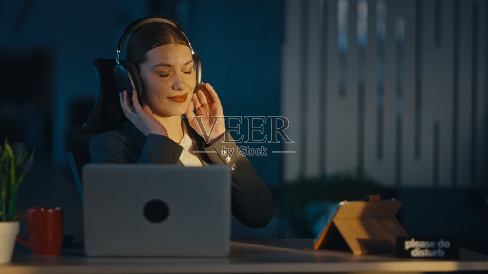 年轻女性穿着商务套装，享受放松时刻，带着宁静的表情通过耳机听音乐。工作与生活平衡、减压、幸福和自我关怀的概念。照片摄影图片