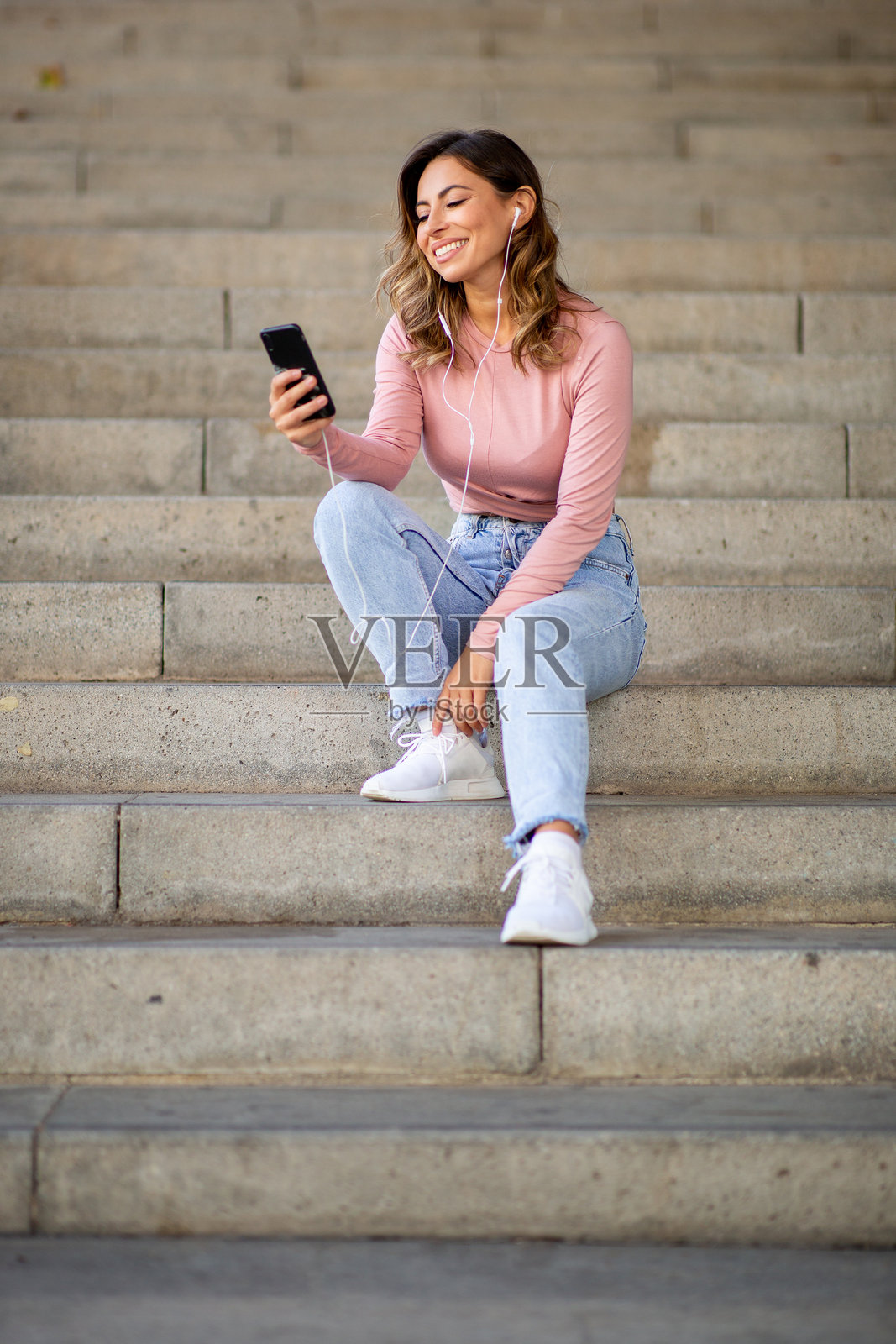 全身快乐的女性坐在楼梯上，手里拿着手机，耳朵里戴着耳机。照片摄影图片