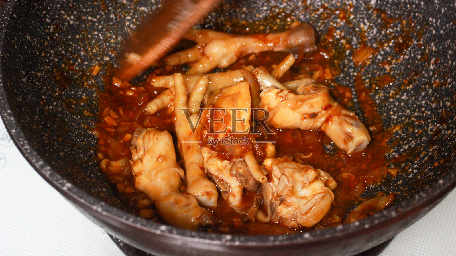 一个装满香辣红烧鸡爪的炒锅，鸡爪在浓郁的美味酱汁中烹饪，旁边有切碎的红辣椒和青辣椒。一把木铲静静地放在锅中，突显出正在进行的家常菜。照片摄影图片