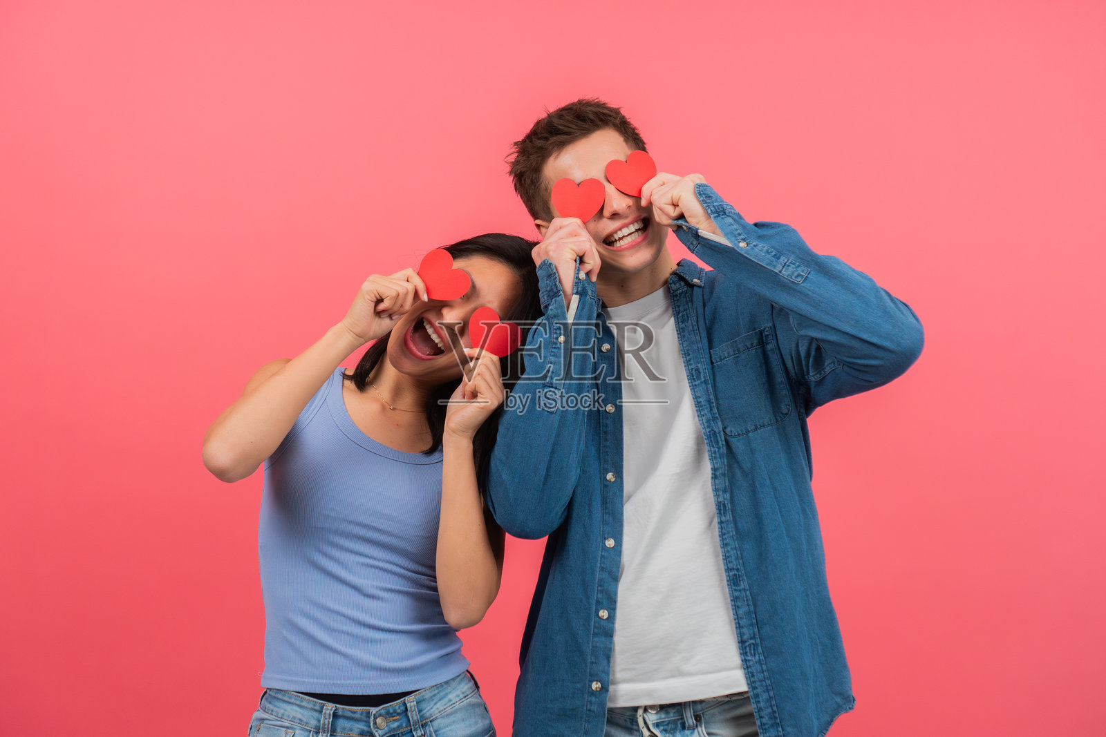 年轻情侣肖像，手持心形物品遮住眼睛，背景为红色。浪漫的男女在情人节庆祝周年纪念，留有空白空间。照片摄影图片