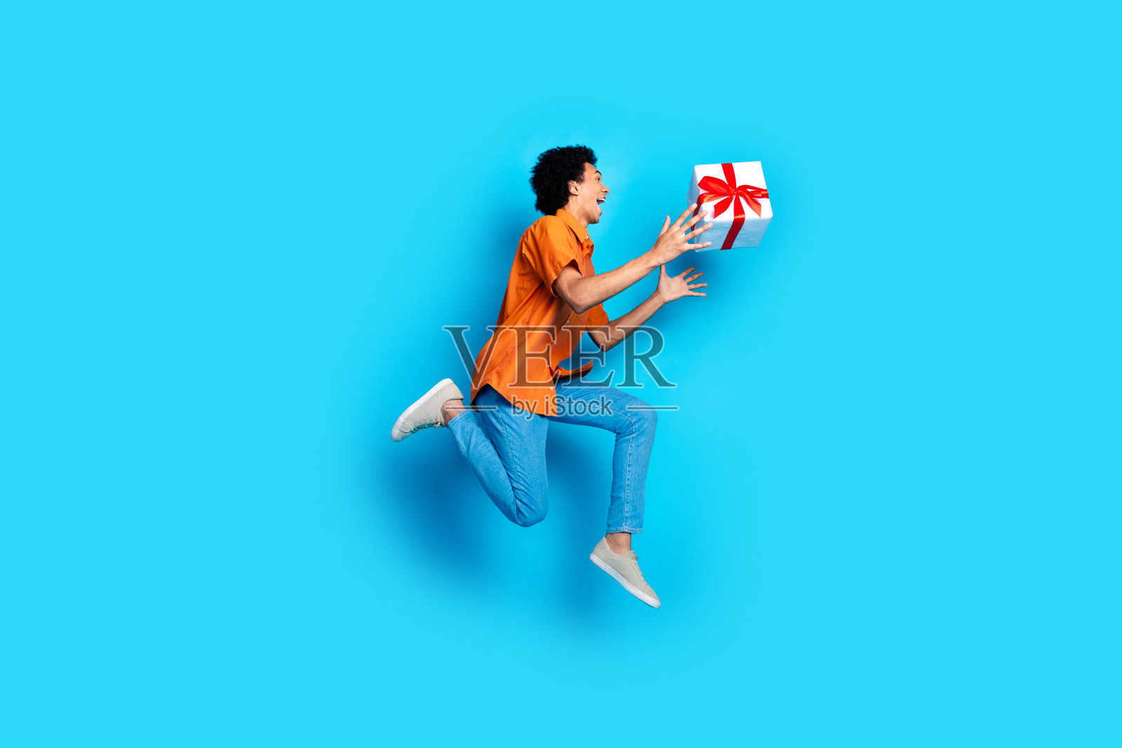 穿着橙色衬衫的快乐积极男士在蓝色背景上高高跳起，手中拿着一个礼物盒，周围留有空白空间。照片摄影图片