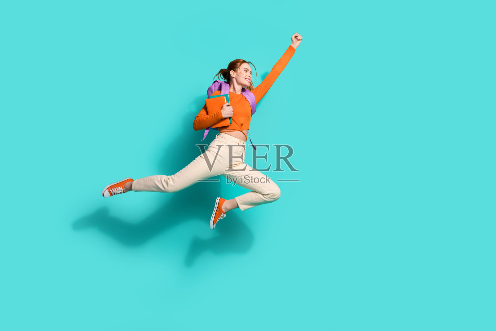 穿着橙色短款上衣的漂亮女性全身照，手持背包和笔记本，飞向空旷的空间，背景为青绿色。照片摄影图片