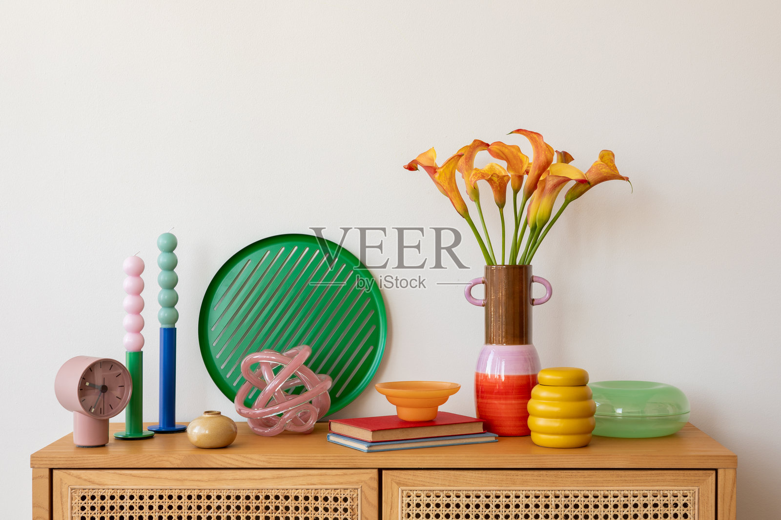 现代桌面装饰，橙色的卡拉百合放在陶瓷花瓶中，五彩缤纷的蜡烛和抽象装饰品摆放在木质橱柜上，营造出一个充满活力和时尚感的室内场景。照片摄影图片